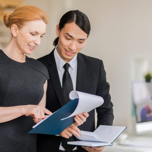 Attractive blond-haired manager with clipboard in hands showing financial figures to young Asian coworker, interior of open plan office on background
